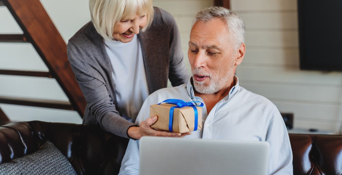 Wife gives her husband gift for the wedding anniversary while he using laptop at home